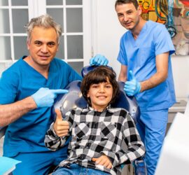 Doi dentiști în uniforme medicale stând lângă un pacient tânăr care zâmbește în scaunul stomatologic.
