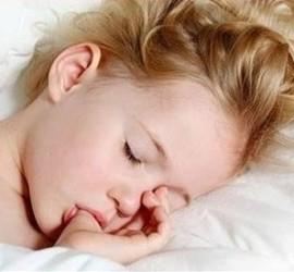 Fetiță cu părul blond, dormind pe o pernă, cu degetul în gură.