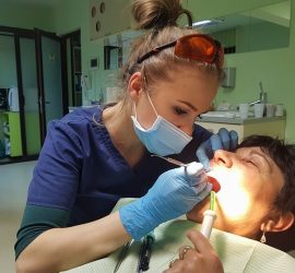 Asistentă dentară cu mănuși și mască examinând gura unui pacient în cabinetul stomatologic.