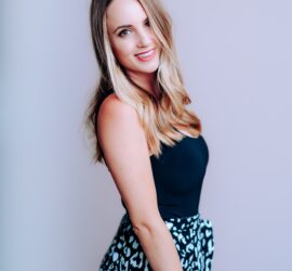 Femeie cu păr lung, îmbrăcată într-o bluză neagră și fustă cu model leopard, zâmbind ușor.