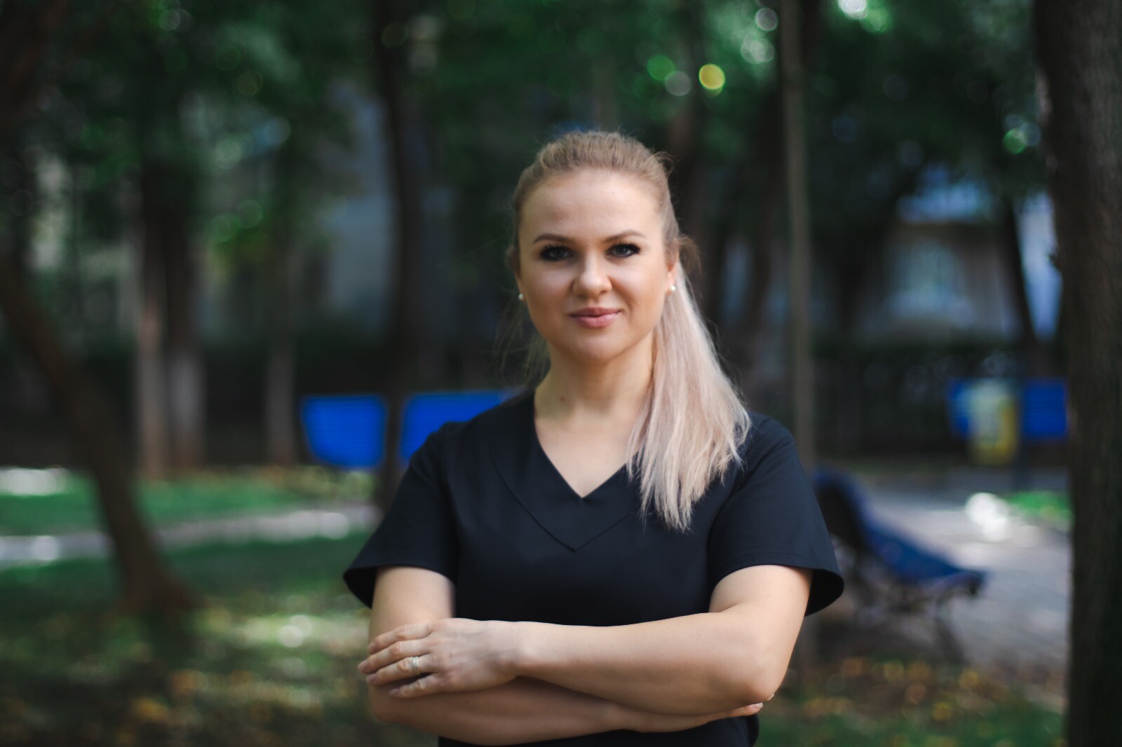 Femeie cu părul lung, îmbrăcată într-o bluză neagră, stând cu brațele încrucișate într-un parc.