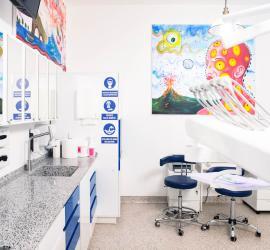 Cabinet dentar modern cu mobilier alb și albastru, dotat cu echipamente dentare și opere de artă colorate pe pereți.