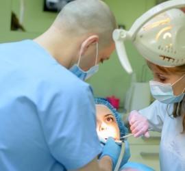 Două persoane în echipamente de protecție efectuează o procedură stomatologică asupra unui pacient.