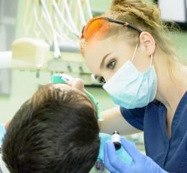 Asistentă stomatologică cu mănuși și mască, examinând un pacient pe un scaun dentar.