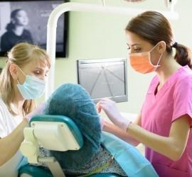 Două asistente dentare îmbrăcate în uniforme, una lucrând la un pacient pe un scaun stomatologic.