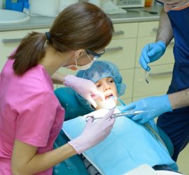 Un copil în scaunul stomatologic, cu un medic dentist și un asistent în timpul unei consultații.