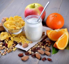 Iaurt în borcan cu lingură, alături de cereale, măr, portocală și nuci pe o masă din lemn.