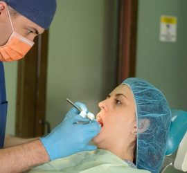 Dentist in blue scrubs and mask examining a patient's mouth in a dental office.