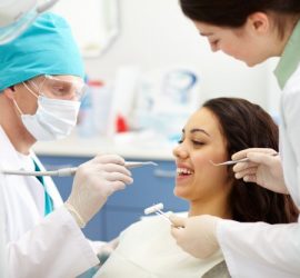 Un dentist examinează un pacient în timp ce o asistentă dentară oferă instrumente dentare.