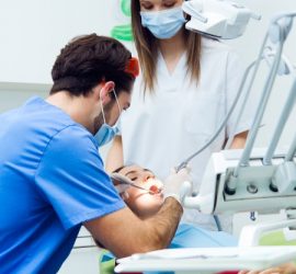 Un dentist examinează un pacient în timpul unei consultații dentare, cu un asistent în fundal.