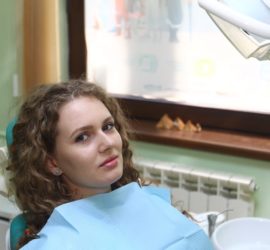 Femeie cu părul ondulat așezată pe un scaun de dentist, purtând un șorț de protecție albastru.