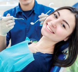 Femeie zâmbitoare așezată pe un scaun de dentist, cu un medic stomatolog în fundal.