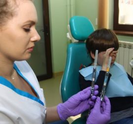 Asistentă dentară arătând două seringi în timp ce un copil stă pe un scaun de dentist.