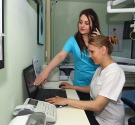 Asistent medical la birou, discutând la telefon și arătând unui coleg pe laptop.