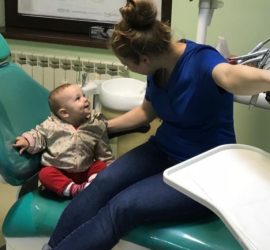 Un copil zâmbitor stând pe un scaun de stomatologie lângă un adult într-un cabinet stomatologic.