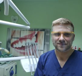 Medic stomatolog purtând o mască de protecție și ochelari în fața unui panou cu imagini dentare.