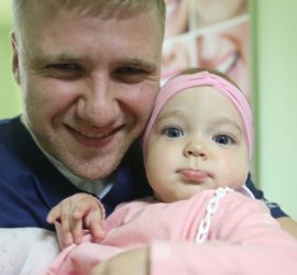 Bărbat zâmbind în timp ce ține un copil mic cu o bentiță roz pe cap.