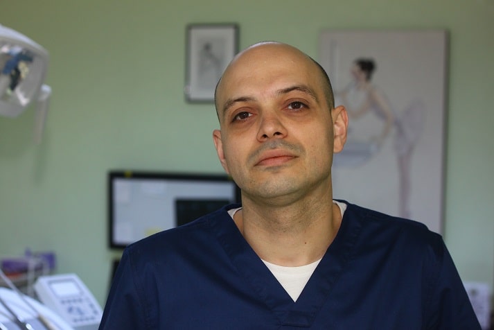 Medic stomatolog cu părul scurt, îmbrăcat în uniformă albastră, privind spre cameră.