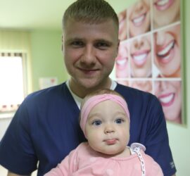 Bărbat îmbrăcat în uniformă medicală ținând un copil mic în brațe, cu un zâmbet pe față.