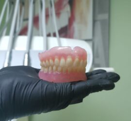 Proteză dentară cu dinți naturali, ținută într-o mână acoperită cu mănușă neagră.