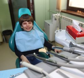 Un copil zâmbitor așezat pe un scaun stomatologic, purtând un șorț de protecție albastru.