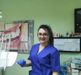 Asistentă dentară zâmbitoare purtând uniformă albastră și mănuși, stând lângă un scaun stomatologic.