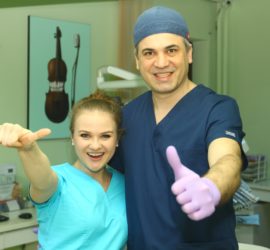 Două persoane în uniformă medicală, zâmbind și arătând cu degetul mare în sus într-o clinică.