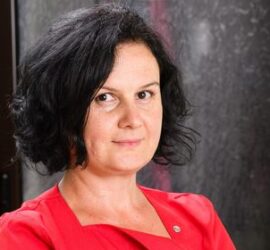 Femeie cu păr negru ondulat, purtând o bluză roșie, zâmbind ușor în fața camerei.