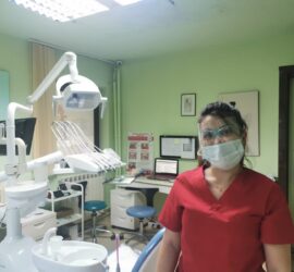 Asistentă medicală stomatologică purtând mască și ochelari de protecție, în fața unui scaun stomatologic.