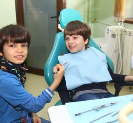 Doi copii în cabinet stomatologic, unul stând pe scaunul dentar și altul ținând un instrument dentar.