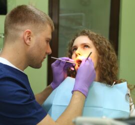 Medic stomatolog cu mănuși și mască examinând gura unei paciente cu părul ondulat.