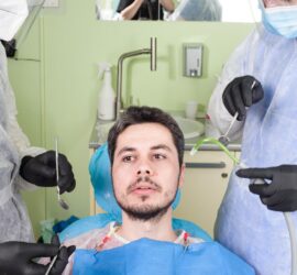 Un pacient așezat pe un scaun de dentist, înconjurat de doi medici stomatologi care folosesc instrumente dentare.