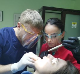 Doi medici dentiști efectuează o procedură stomatologică pe un pacient, purtând echipamente de protecție.