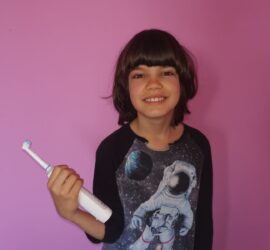 Un copil zâmbind și ținând o periuță de dinți electrică în mână, purtând un tricou cu un astronaut.