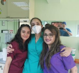 Trei asistente medicale zâmbind în fața unei oglinzi, una dintre ele purtând mască de protecție.