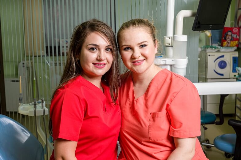 Două asistente medicale zâmbind, îmbrăcate în uniforme roșii și portocalii, într-un cabinet stomatologic.