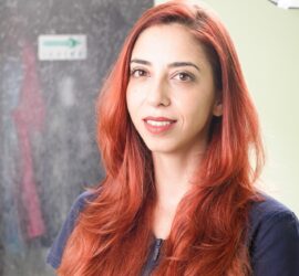 Femeie cu păr lung și roșcat, zâmbind în fața camerei, cu un fundal neclar.