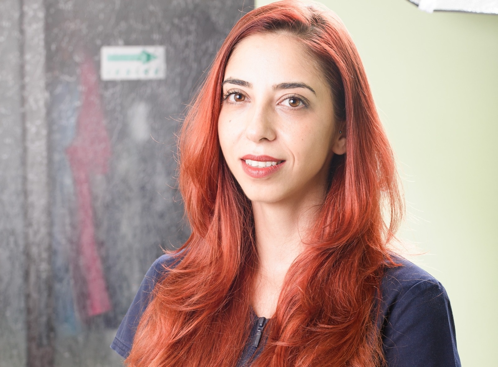 Femeie cu păr lung și roșcat, zâmbind în fața camerei, cu un fundal neclar.