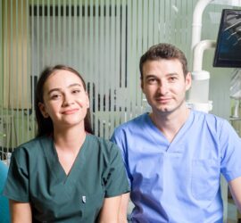Două persoane, un bărbat și o femeie, îmbrăcați în uniforme medicale, zâmbind în fața camerei într-o clinică dentară.