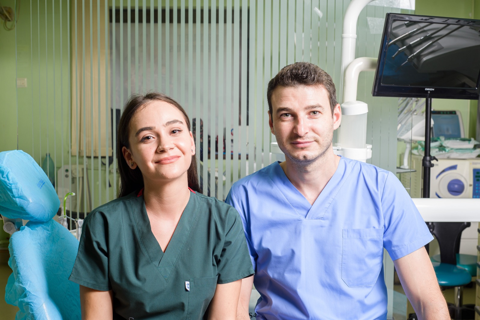 Două persoane, un bărbat și o femeie, îmbrăcați în uniforme medicale, zâmbind în fața camerei într-o clinică dentară.