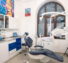 Cabinet stomatologic luminos cu mobilier alb si sertare albastre, scaun dentar albastru si echipamente moderne, fereastra arcuita, iluminare adecvată si detartraj dentar utilizat in clinică.