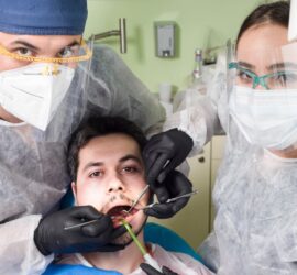 Două persoane îmbrăcate în echipament de protecție efectuează o procedură stomatologică asupra unui pacient.