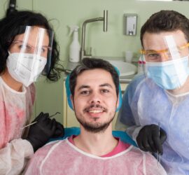 Un pacient zâmbind în timp ce este însoțit de doi medici dentiști, purtând echipament de protecție.