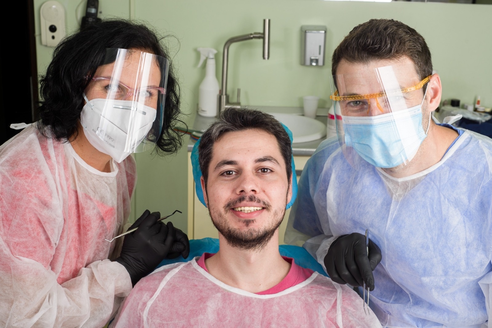 Un pacient zâmbind în timp ce este însoțit de doi medici dentiști, purtând echipament de protecție.