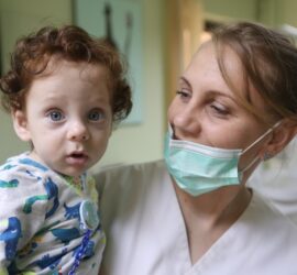 Medic cu mască care ține un copil cu părul creț și ochi deschiși în brațe.