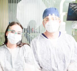 Două persoane în echipament de protecție medicală, cu măști și viziere, într-un cabinet medical.