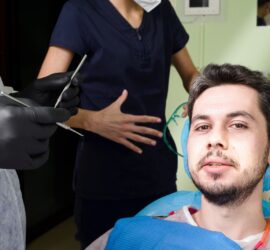 Pacient zâmbind în timp ce se află pe un scaun stomatologic, cu personal medical în fundal.