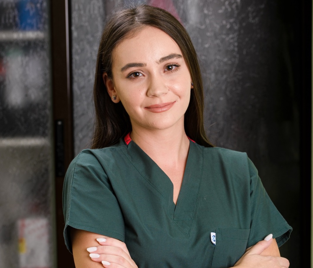 Asistent medical tânăr zâmbind, îmbrăcat în uniformă verde, cu brațele încrucișate.