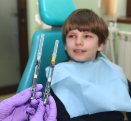 Un copil zâmbind în scaunul de dentist, ținând două seringi în mână.