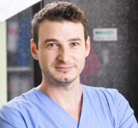 Bărbat cu părul brun, îmbrăcat în halat medical albastru, zâmbind în fața camerei.